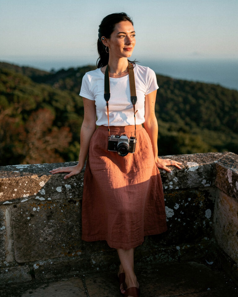 Woman standing on the stone ramparts of the Moorish Castle in Sintra, warm side-light on her face, forested hills and faint ocean horizon behind her.