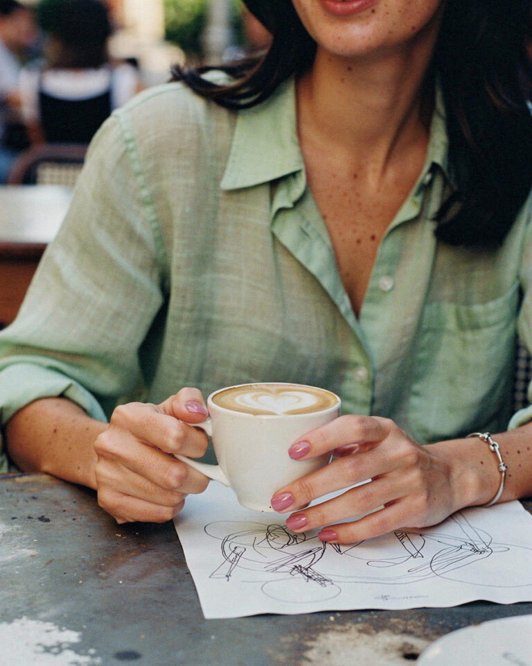 Woman sitting alone at an outdoor café terrace near the Metropol Parasol in Seville, sketching on a napkin beside a café con leche, warm early morning light.