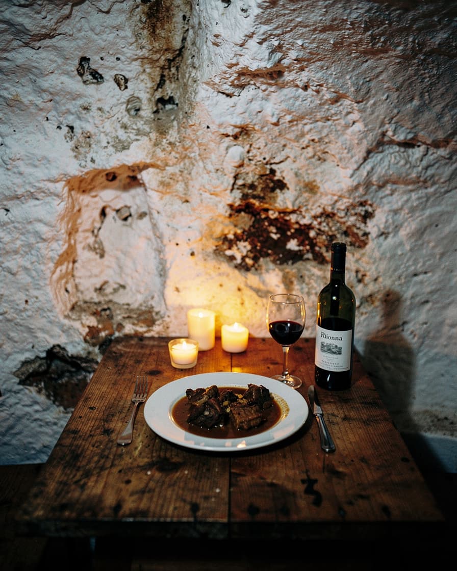 Plate of oxtail stew and a glass of red wine on a rough wooden table inside a whitewashed cave restaurant, warm candlelight on the walls.