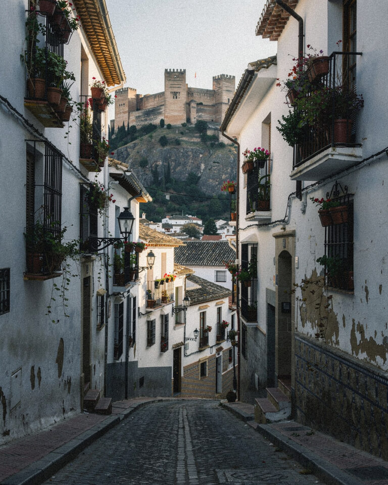 Steep cobblestone street in Granada's Albaicín quarter lined with whitewashed houses and flowering balconies, Alhambra visible on the opposite hill in warm morning light.