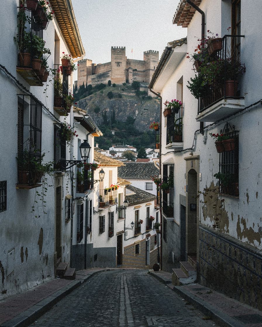 Steep cobblestone street in Granada's Albaicín quarter lined with whitewashed houses and flowering balconies, Alhambra visible on the opposite hill in warm morning light.
