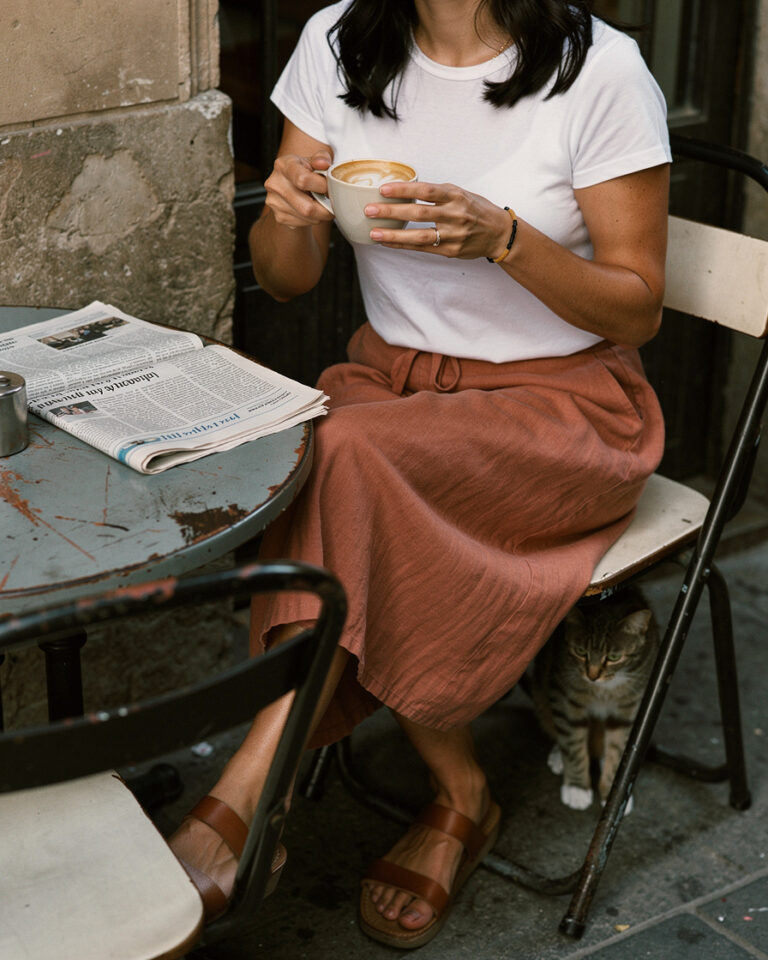 Sunlit terrace table in Barcelona's Gràcia neighborhood with a milky coffee and open newspaper, warm early morning light across stone paving.