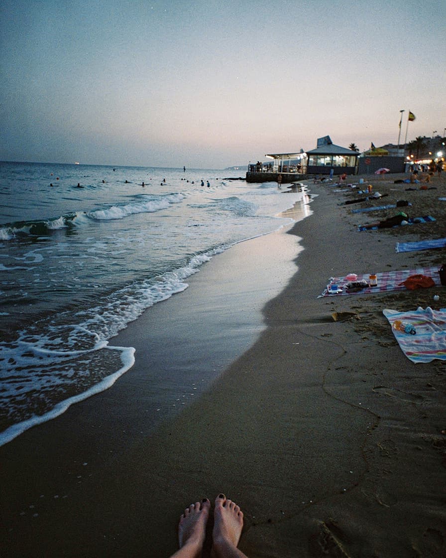 Empty stretch of Barceloneta beach at dusk with wet sand reflecting fading pink light, a pair of shoes and a film camera in the foreground.