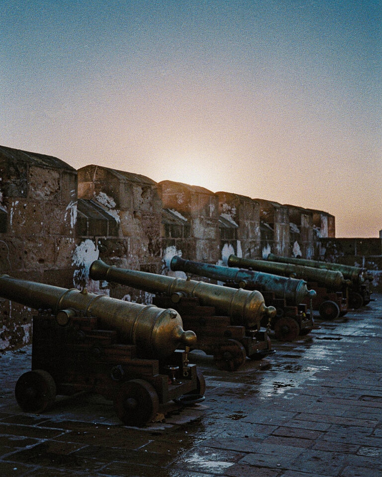 Silhouetted brass cannons along the Skala ramparts of Essaouira at sunset, ocean spray catching warm light against dark stone, no people in frame.