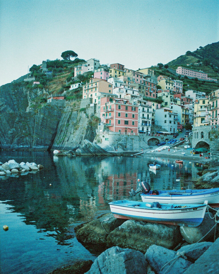 manarola cinque terre dawn: Pastel-colored stacked houses of Manarola reflected in calm harbor water at dawn, fishing boats o