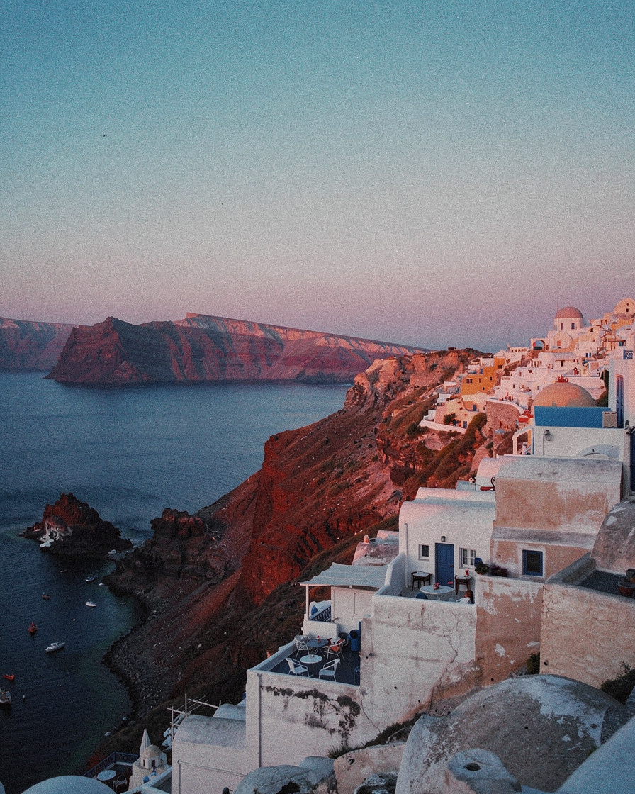 santorini sunset film photography: Whitewashed buildings of Santorini clinging to caldera cliffs glowing pink-gold at sunset,