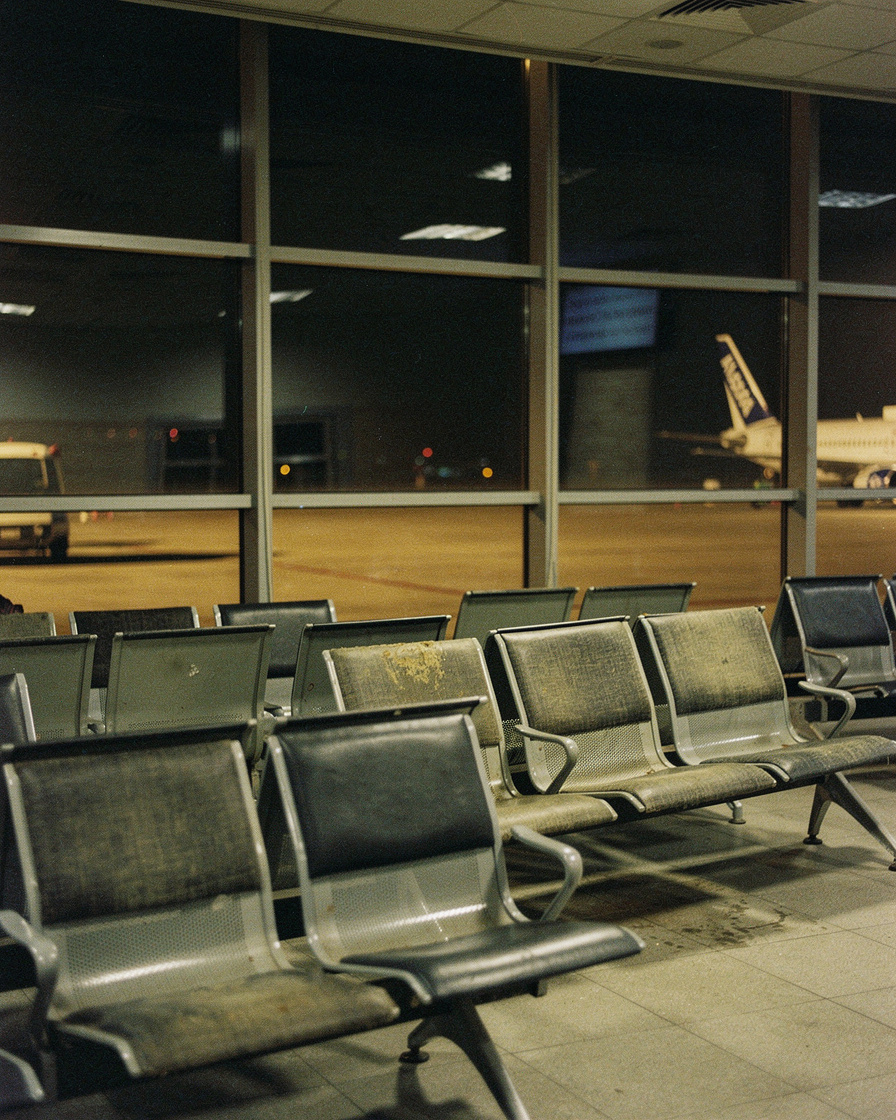 tbilisi airport night layover: Empty airport gate seating under flat ceiling light, dark apron beyond glass with one illumina
